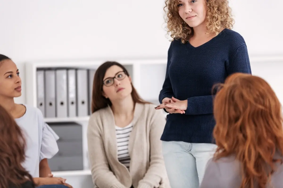 A diverse group of women holding coffee cups, sitting in a circle in a sunlit living room for a group therapy session